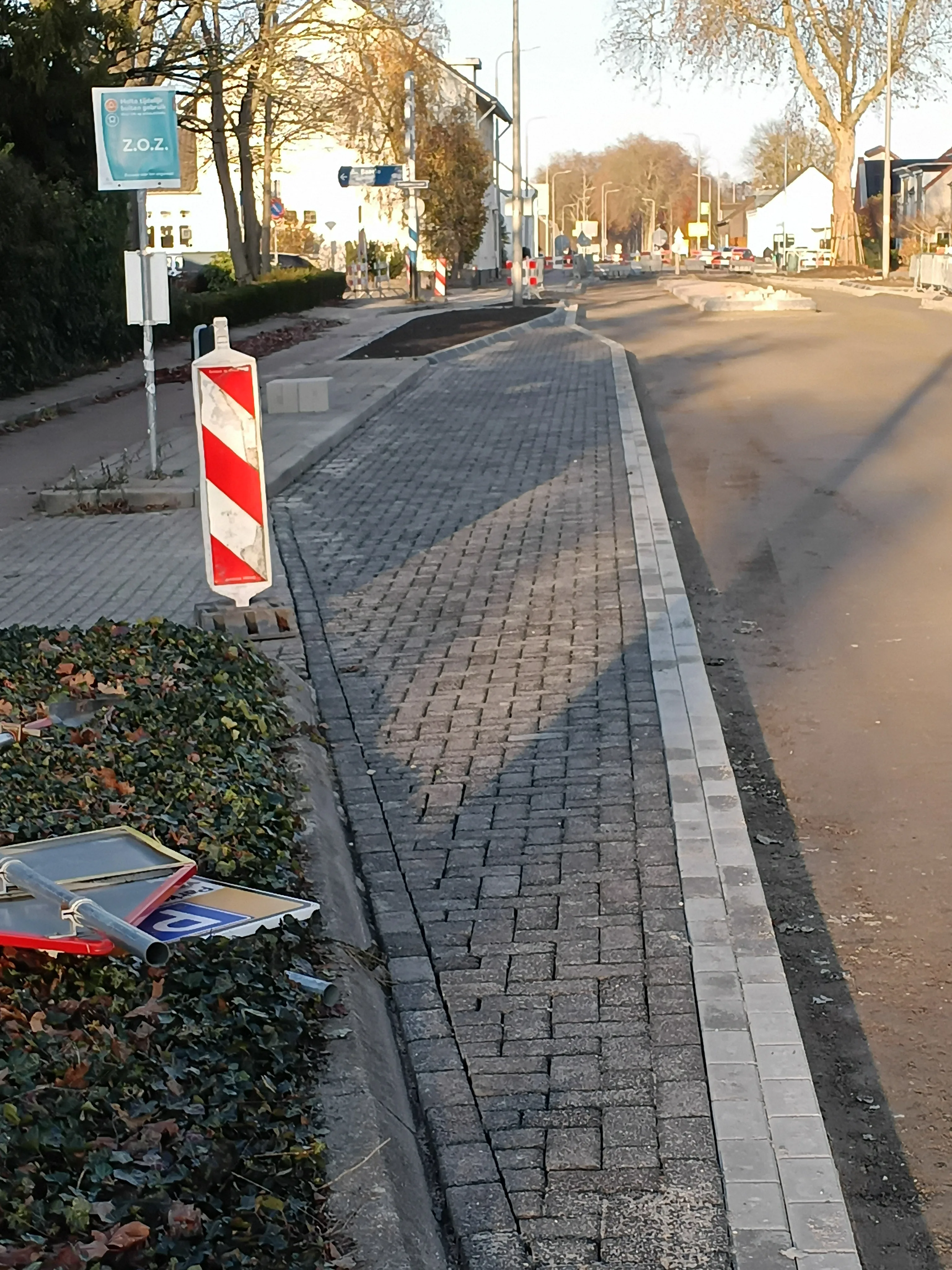 Nieuw aangelegde trottoir met grijze klinkerstenen en lichte opsluitbanden, gemarkeerd door rood-witte werkblokkering